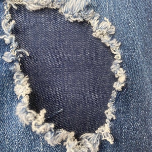 Pu&ograve; includere: Primo piano di un tessuto denim blu con un buco al centro. Il buco &egrave; circondato da bordi sfilacciati.