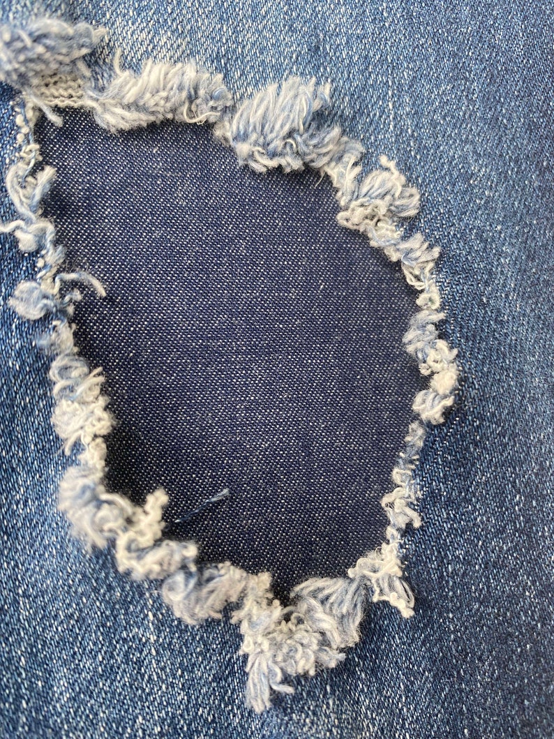 Pu&ograve; includere: Primo piano di un tessuto denim blu con un buco al centro. Il buco &egrave; circondato da bordi sfilacciati.