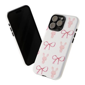 May include: A white phone case featuring a pink bunny rabbit and bow design. The case has a black camera bump and a black border.
