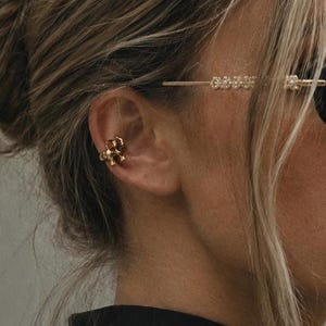 Könnte beinhalten: Ein goldfarbener Ohrklemmer mit einer kleinen, detaillierten Elefantenfigur. Das metallische Accessoire ist am äußeren Knorpel des Ohrs angebracht. Das Design ist poliert und reflektierend und zeigt die deutliche Form des Rüssels und der Ohren.