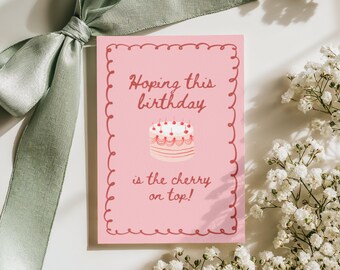 May include: A pink birthday card with a scalloped border and the text "Hoping this birthday is the cherry on top!" A cake with cherries is illustrated on the card. A sage green ribbon and white flowers are in the background.