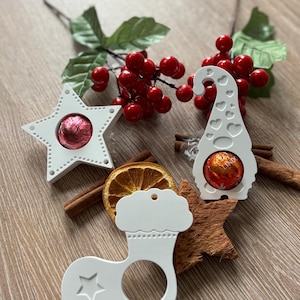 May include: White Christmas decorations in star, gnome, and stocking designs, each holding a chocolate. Cinnamon sticks, dried orange slices, and red berries provide festive touches. The items are arranged on a wooden surface.
