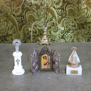 May include: Three silver religious figurines on white marble bases. The center figurine is a framed image of a man in a robe. The figurine on the left is a woman with a halo. The figurine on the right is a woman wearing a white robe with a gold crown.