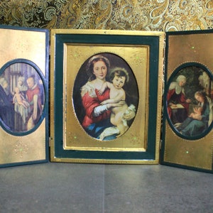 May include: A three-panel wooden triptych with gold leaf accents. Each panel features a framed oval image of a religious scene. The center panel depicts the Virgin Mary holding the baby Jesus. The left panel shows a scene of the Nativity, and the right panel shows a scene of the Annunciation.