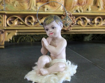 May include: A small, white figurine of a child with a halo, sitting on a white fluffy base. The child is wearing a white cloth and has its hands clasped in prayer. The figurine is in front of a wooden, ornate altar.
