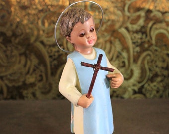 May include: A small figurine of a child holding a wooden cross. The child is wearing a blue and white robe and has a halo above their head.