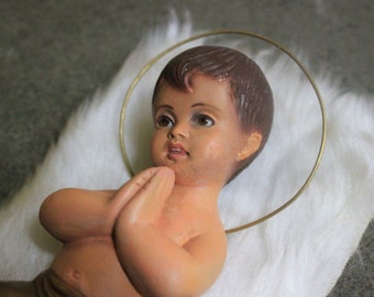 May include: A vintage figurine of a baby Jesus lying on a white faux fur surface. The figurine is made of painted plaster and has a gold halo.