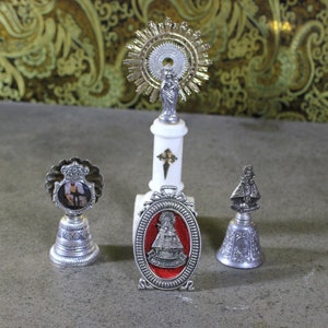 Könnte beinhalten: Vier silberne religiöse Glocken mit Bildern von Heiligen und einem Kreuz. Die Glocken stehen auf einer weißen Oberfläche.
