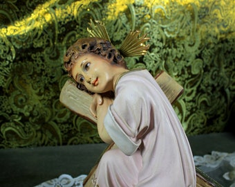 May include: A white porcelain figurine of a young angel with a halo, resting their head on a wooden cross. The angel is wearing a white robe and has a soft, delicate expression.