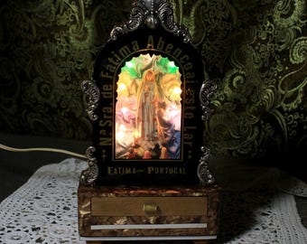 Può includere: Un santuario religioso decorativo con una figura di santa illuminata da luci colorate. La cornice nera ha dettagli argentati e il testo "N. Sra. de Fátima Abençoe este Lar" e "Fátima - Portugal". Include un piccolo cassetto.