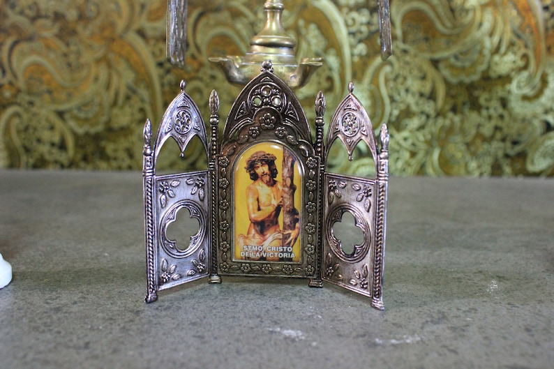 May include: A silver-colored, ornate, three-panel display stand with a small image of a man with a crown of thorns. The image is labeled "SEMI CRISTO DELA VICTORIA".