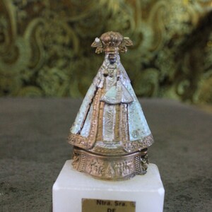 May include: A small, gold-colored statue of the Virgin Mary, wearing a blue and white robe, on a white marble base. The base has a gold plaque with the text "Ntra. Sra. DE GUADALUPE".