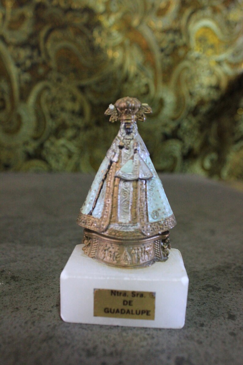 May include: A small, gold-colored statue of the Virgin Mary, wearing a blue and white robe, on a white marble base. The base has a gold plaque with the text "Ntra. Sra. DE GUADALUPE".