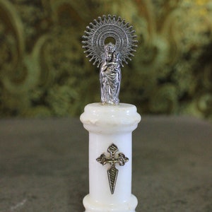 May include: A white marble statue of the Virgin Mary and baby Jesus with a silver halo and cross. The statue is on a white marble base with the inscription "N.S. del Pilar".