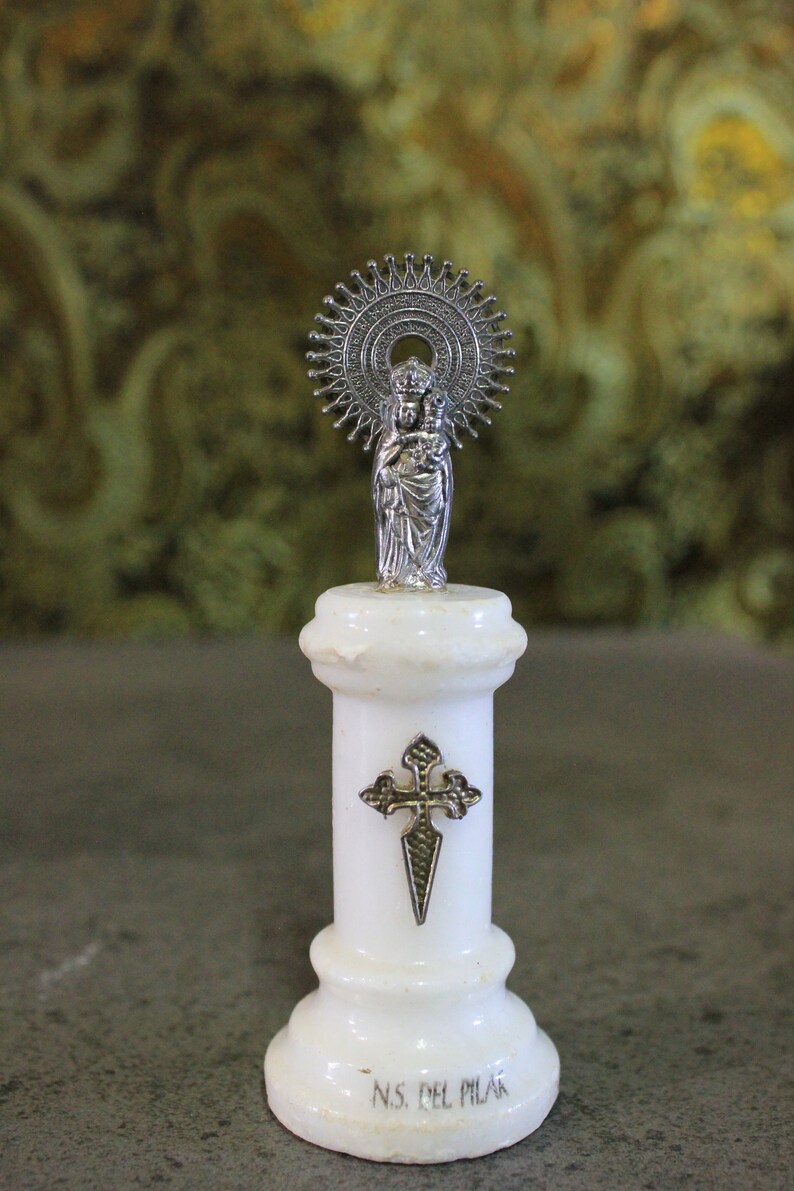 May include: A white marble statue of the Virgin Mary and baby Jesus with a silver halo and cross. The statue is on a white marble base with the inscription "N.S. del Pilar".