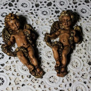 Puede incluir: Dos figuras de querubines antiguos con alas detalladas y guirnaldas florales. Los querubines están pintados en tonos marrones y dorados, y están sobre una superficie de encaje blanco. Estas figuras decorativas son ideales para la decoración del hogar.