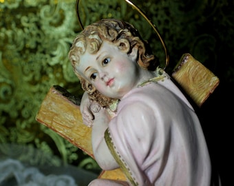 May include: A figurine of a child with a halo, wearing a pink robe, holding a wooden cross. The figurine is made of painted porcelain and has a realistic appearance.