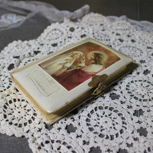 May include: A small, vintage religious book with a gold clasp, featuring an image of Jesus and a woman in a white veil. The book is set on a white lace doily, with a gray background.