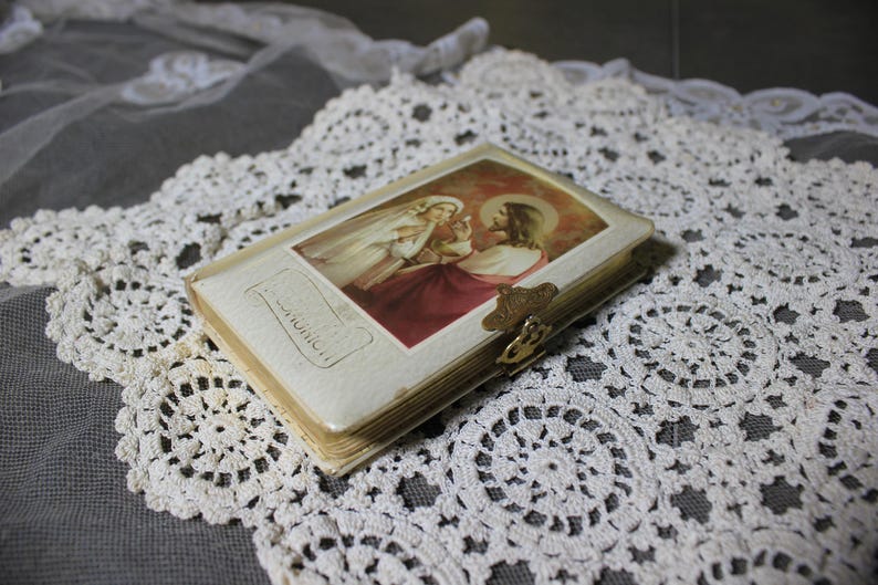 May include: A small, vintage religious book with a gold clasp, featuring an image of Jesus and a woman in a white veil. The book is set on a white lace doily, with a gray background.