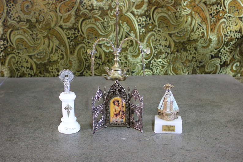 May include: Three silver religious figurines on white marble bases. The center figurine is a framed image of a man in a robe. The figurine on the left is a woman with a halo. The figurine on the right is a woman wearing a white robe with a gold crown.