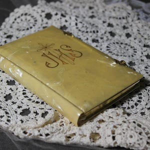 May include: A vintage, rectangular, cream-colored book-shaped case with gold trim and a clasp. The cover features gold lettering, including the letters "JHS". The case rests on a white lace doily.