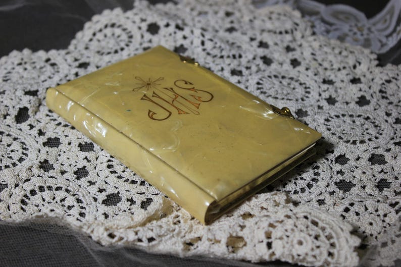 May include: A vintage, rectangular, cream-colored book-shaped case with gold trim and a clasp. The cover features gold lettering, including the letters "JHS". The case rests on a white lace doily.