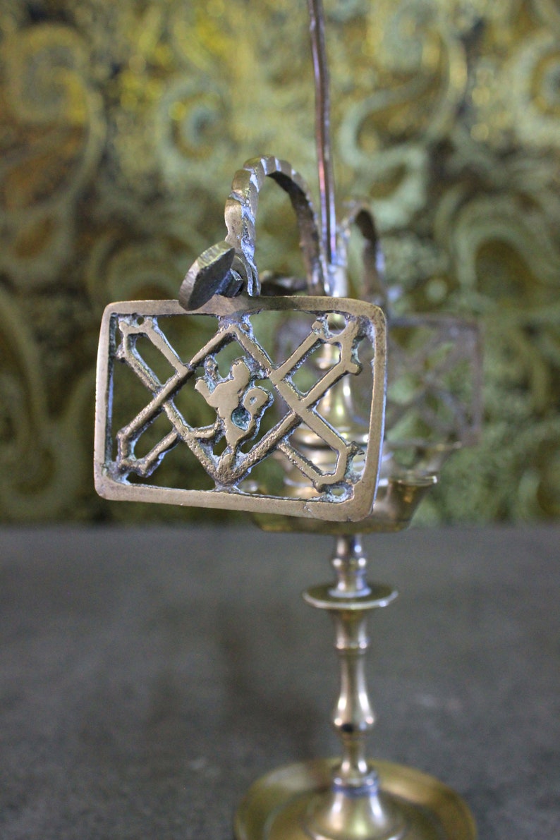 May include: A brass candlestick with a decorative design featuring a cross and a figure. The candlestick has a round base and a tall, slender stem.