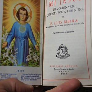 May include: Open book with a religious illustration of Jesus in a blue robe, surrounded by white lilies and a golden halo. Text in Spanish reads "Mi Jes&uacute;s" and "Devocionario". Published in Madrid, 1954.