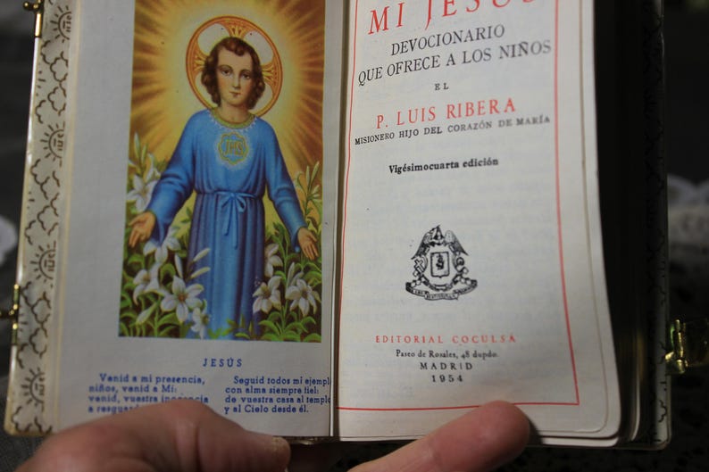 May include: Open book with a religious illustration of Jesus in a blue robe, surrounded by white lilies and a golden halo. Text in Spanish reads "Mi Jes&uacute;s" and "Devocionario". Published in Madrid, 1954.