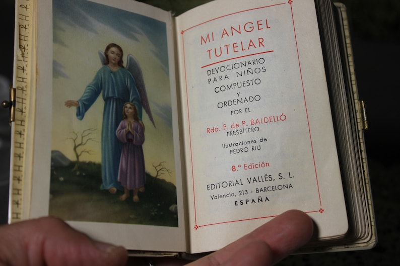 May include: Open book with the title "MI ANGEL TUTELAR" in Spanish. The left page features an illustration of an angel and a child. The right page has text in Spanish, including the author's name and the publisher's address.