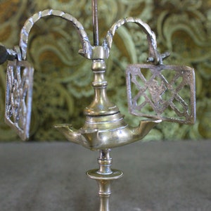 May include: A brass oil lamp with three arms and a decorative design. The lamp has a central stem with a round base and a decorative top. The arms are curved and have a decorative design. The lamp is on a wooden surface.