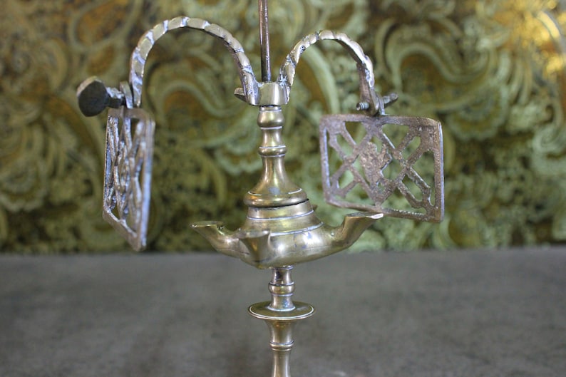 May include: A brass oil lamp with three arms and a decorative design. The lamp has a central stem with a round base and a decorative top. The arms are curved and have a decorative design. The lamp is on a wooden surface.