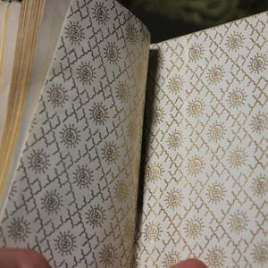 May include: Open journal with gold-colored clasp and pages. The interior lining features a repeating pattern of gold sunbursts and geometric shapes on a cream background. The pages are a light tan color.
