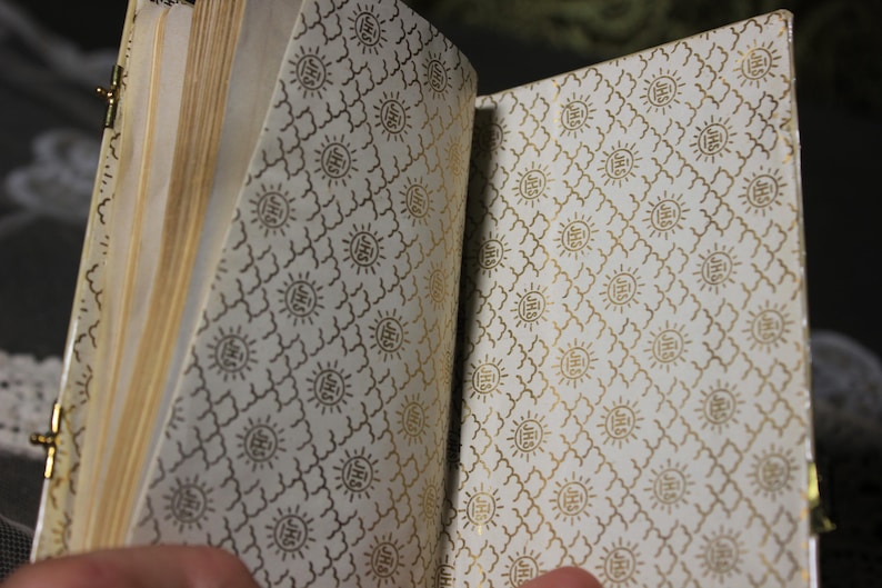 May include: Open journal with gold-colored clasp and pages. The interior lining features a repeating pattern of gold sunbursts and geometric shapes on a cream background. The pages are a light tan color.