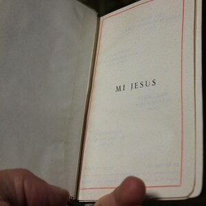 May include: An open book with the words "MI JESUS" printed on the right page. The pages are cream-colored with a red border. The book has a decorative gold clasp and a patterned spine.