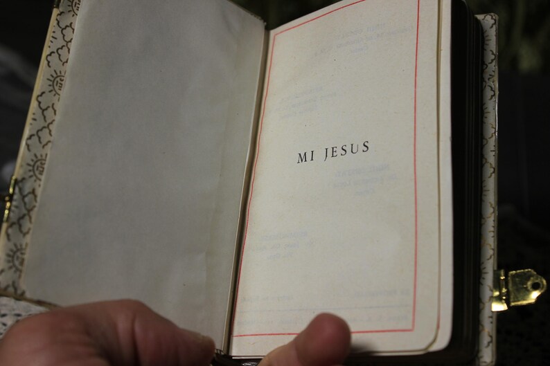 May include: An open book with the words "MI JESUS" printed on the right page. The pages are cream-colored with a red border. The book has a decorative gold clasp and a patterned spine.