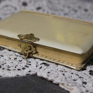 May include: A vintage cream-colored book with a decorative gold clasp. The book's pages are aged, and the cover is slightly worn. The book is resting on a white lace doily, creating a classic, antique aesthetic.