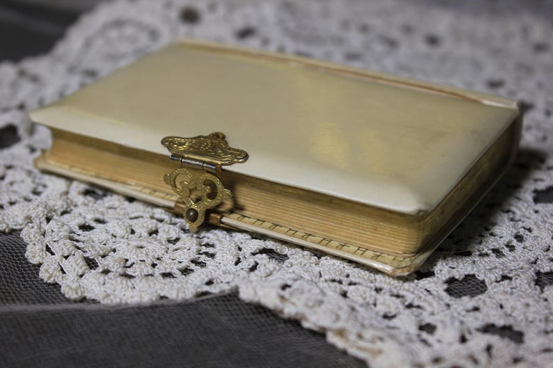 May include: A vintage cream-colored book with a decorative gold clasp. The book's pages are aged, and the cover is slightly worn. The book is resting on a white lace doily, creating a classic, antique aesthetic.