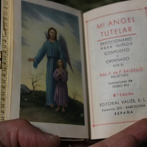 May include: Open book with a color illustration of an angel and a child. The text on the right page reads "MI ANGEL TUTELAR" and details about the book's author and publisher. The book is held open.