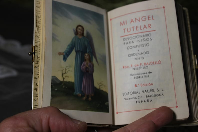 May include: Open book with a color illustration of an angel and a child. The text on the right page reads "MI ANGEL TUTELAR" and details about the book's author and publisher. The book is held open.