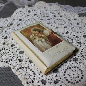 May include: A small, off-white book with a religious image on the cover, featuring a woman in a white veil and a man in red robes. The book is set on a white lace doily with a floral pattern. The word "COMMUNION" is visible.