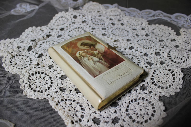 May include: A small, off-white book with a religious image on the cover, featuring a woman in a white veil and a man in red robes. The book is set on a white lace doily with a floral pattern. The word "COMMUNION" is visible.