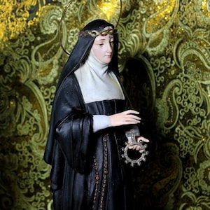 May include: A statue of a woman in a black habit and white wimple, holding a rosary and a decorative ring. She wears a crown of thorns. The background is a gold and green patterned fabric.