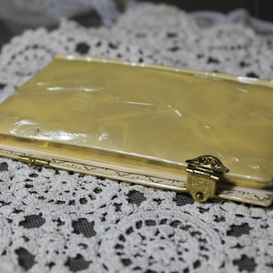 May include: A vintage, rectangular, cream-colored compact with a gold clasp. The compact has a pearlescent, marbled finish on the lid and is resting on a white lace doily. The clasp is ornate and gold-toned.