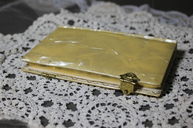 May include: A vintage, rectangular, cream-colored compact with a gold clasp. The compact has a pearlescent, marbled finish on the lid and is resting on a white lace doily. The clasp is ornate and gold-toned.