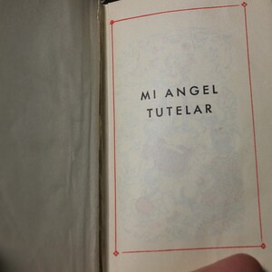 May include: Open book with the text "MI ANGEL TUTELAR" in black letters on a cream-colored page. The text is inside a red border. The book is open to the page with the text.