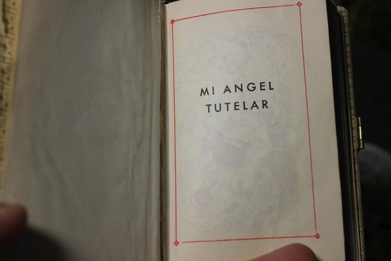 May include: Open book with the text "MI ANGEL TUTELAR" in black letters on a cream-colored page. The text is inside a red border. The book is open to the page with the text.