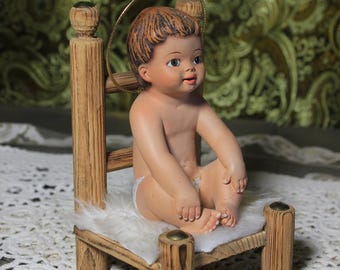 May include: A ceramic figurine of a baby with brown hair and a gold halo, seated on a small wooden bed with a white fur-like cushion. The bed has four wooden posts and a wooden headboard. The baby is wearing a diaper.