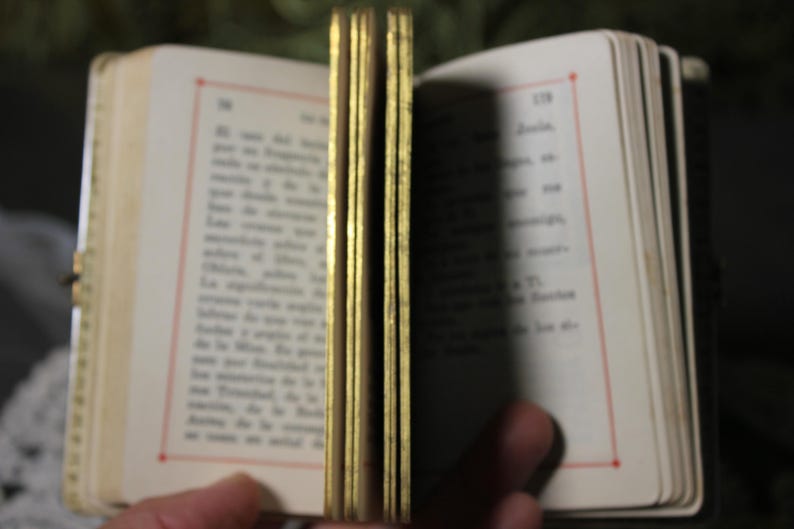 May include: An open antique book with gold-gilded edges. The pages are cream-colored with printed text and red border lines. The book is held open, revealing the text and the gold edges.