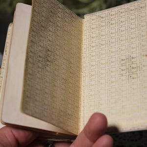 May include: A small, vintage notebook with a cream-colored cover and pages. The interior pages feature a repeating gold pattern. The notebook is open, revealing the patterned interior. A gold clasp secures the notebook.